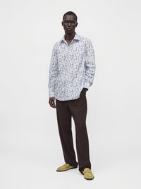 Homme portant une chemise blanche à manches longues avec un imprimé floral bleu, un pantalon brun foncé et des mocassins jaunes, debout devant un fond clair uni.