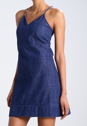 Mujer vestida con un vestido sin mangas de mezclilla azul oscuro, con tirantes delgados, escote en V y falda evasé frente a un fondo claro.