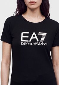 Γυναίκα που φορεί μαύρο μπλουζάκι με λευκό λογότυπο EA7 Emporio Armani στο κέντρο του στήθους, κοντά μανίκια και στρογγυλή λαιμόκοψη.