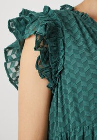 Τιρκουάζ μπλούζα με ρυτιδωτά μανίκια, υφασματική υφή σε ζιγκ-ζαγκ σχέδιο και ημιδιαφανές ύφασμα. Διαθέτει λεπτές περιλαβές, αναδεικνύοντας τόσο το ύφασμα όσο και το σχέδιο.