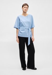 Ανοιχτό μπλε τοπ κοντό μανίκι με ζώνη στο ύψος της μέσης, σε συνδυασμό με μαύρους παντελόνες φαρδιάς γραμμής. Απαλή ύφανση, μινιμαλιστικός σχεδιασμός και χαλαρή εφαρμογή.