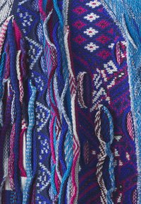 Adīta pončo z zilas, purpura un baltas krāsas ar sarežģītām ģeometriskām rakstām, ar frizētām malām un dažādām teksturētām šķiedrām visā garumā.