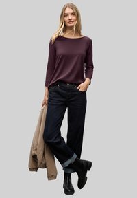 Langarm, diep paarse top gecombineerd met donkere denim jeans, opgerolde mouwen en zwarte enkellaarzen. Beige jas over één arm gedrapeerd.