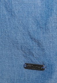 Vaate denim kangas vaaleansinisena, jossa on sileä pinta. Varustettu pienellä mustalla metallilapulla, jossa on painettu "Denim". Huomaamaton ompelukuvio on näkyvissä.