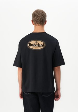 Black oversized t-shirt made of cotton, featuring a circular beige Timberland logo on the back with "ACTIVE RUGGED GEAR" text underneath.