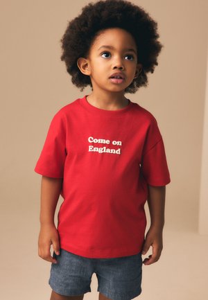 Jeune enfant aux cheveux bouclés portant un t-shirt rouge avec le texte "Come on England" et un short bleu, regardant légèrement vers le haut sur un fond uni.