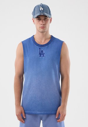 Hombre con camiseta azul sin mangas y gorra de béisbol a juego desgastada con logo blanco de "LA", de pie frente a un fondo blanco.