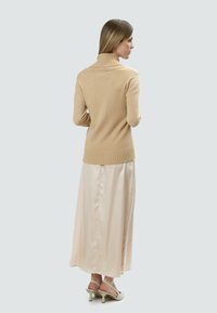 Beiger Strickpullover mit hohem Kragen und geripptem Saum, kombiniert mit einem weichen, satinartigen, hellrosa Rock. Dazu werden hellfarbige, hochhackige Schuhe getragen.