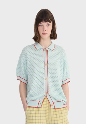 Mujer con un cárdigan de punto azul claro con ribetes y botones rojos, combinando con pantalones de cuadros amarillos, posando frente a un fondo liso.