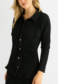 Black pleated blouse with a collar, button front, and tie waist. Features gold buttons and long sleeves with textured fabric.