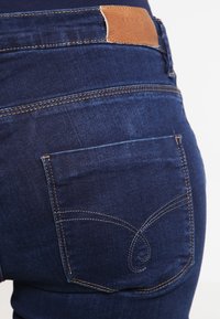 Tmavě modré džíny z denimu s hladkou strukturou, s hnědou koženou nášivkou a detailním prošíváním na zadních kapsách.