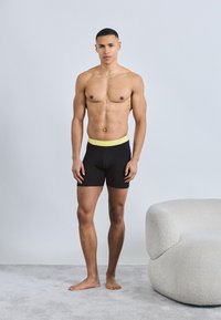 Μυώδης άνδρας που στέκεται ξυπόλητος σε χαλί, φορώντας μαύρα μποξεράκια με κίτρινη μέση, δίπλα σε μοντέρνο λευκό επενδεδυμένο καρέκλα.