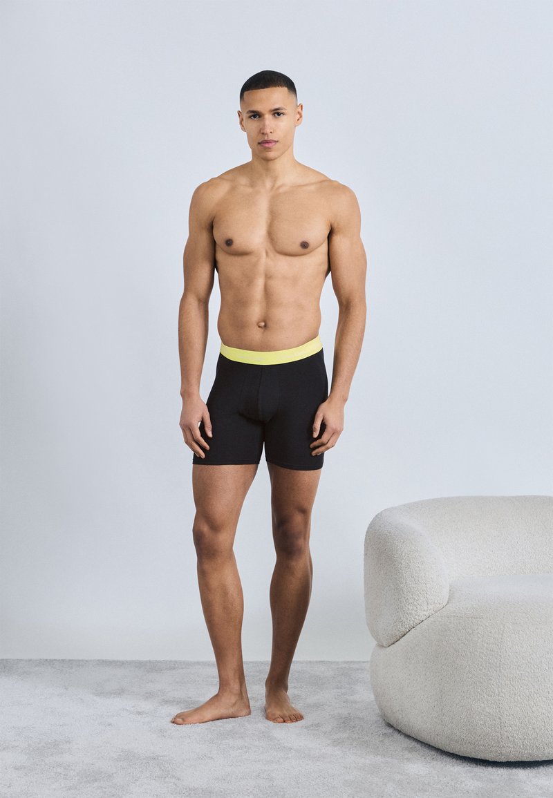 Μυώδης άνδρας που στέκεται ξυπόλητος σε χαλί, φορώντας μαύρα μποξεράκια με κίτρινη μέση, δίπλα σε μοντέρνο λευκό επενδεδυμένο καρέκλα.