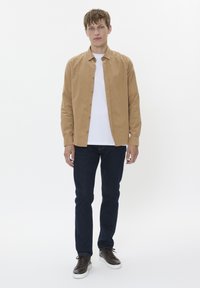 Een tan button-up shirt over een wit T-shirt, gecombineerd met donkerblauwe jeans en bruine sneakers. Eenvoudig ontwerp, standaard pasvorm en een gladde textuur.