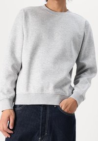 Sweat-shirt en coton gris avec un col rond, des poignets et un ourlet côtelés. Présente une texture douce et lisse ainsi qu'une coupe décontractée. Porté avec un jean foncé.