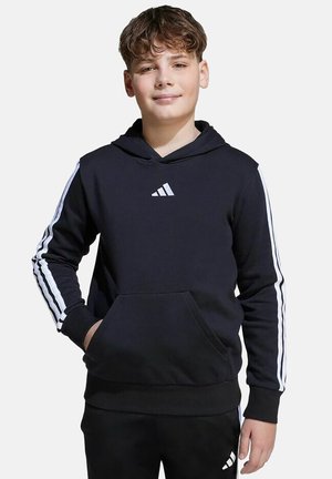 Jongen draagt een zwarte Adidas-hoodie en bijpassende zwarte broek met witte strepen, staat met één hand in de zak en glimlacht licht.