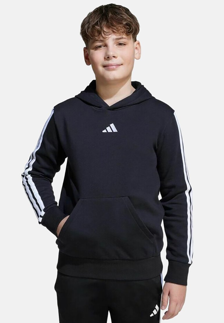 Garçon portant un sweat à capuche Adidas noir et un pantalon noir assorti avec des bandes blanches, debout avec une main dans la poche, souriant légèrement.