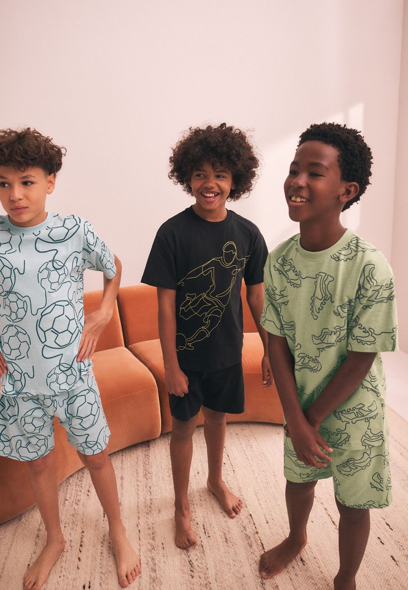Tre barefodede drenge står indendørs iført matchende mønstrede t-shirts og shorts med fodboldtema, smilende og afslappede.