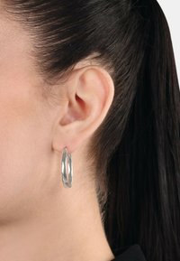 Silverfärgade ringörhängen med en slank, förlängd form och polerad yta, visade på en modells öra mot mörkt hår.