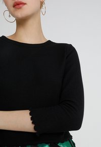Top negro de manga larga con canalé, escote redondo y dobladillo ondulado, combinado con una falda verde estampada y grandes pendientes de aro dorados.