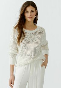 Weißer gestrickter Pullover mit einem strukturierten Muster und gerippten Bündchen. Kombiniert mit hellen Hosen für einen kontrastierenden Look.