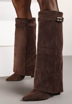 Bottes marron en daim jusqu'au genou avec des bouts pointus, une fermeture éclair sur le côté et une sangle décorative avec boucle près du haut.