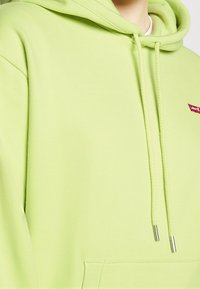 Sudadera verde lima con capucha ajustable, puños acanalados y un bolsillo frontal. Presenta un pequeño logo rojo de Levi's en el pecho. Textura suave.
