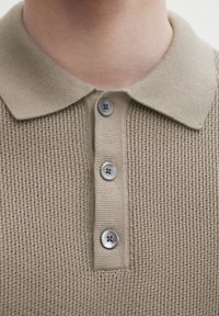 Beige neulottu poolopaita, jossa on teksturoitu kangas, tasainen kaulus ja kolme harmaata nappia edessä kiinnitystä varten.