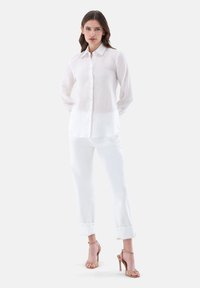 Camicia button-up bianca in lino con maniche lunghe, abbinata a pantaloni bianchi e sandali con tacco beige su uno sfondo neutro.