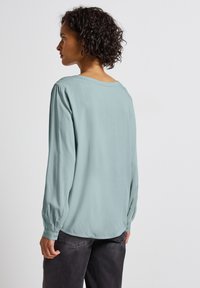 Blouse bleu clair à manches longues, coupe décontractée, encolure ronde et accents de boutons aux poignets. Texture lisse, léger éclat.