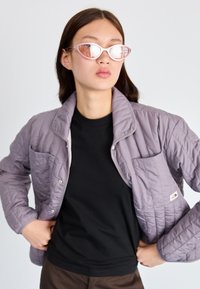 Quiltet pastellilla lilla jakke med to frontlommer, kombinert med en svart skjorte. Modellen har på seg lyserosa kattøye-solbriller.