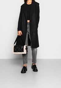 Černý bezrukávový kabát přes černý žebrovaný krátký top, šedé džíny a černé platformové mokasíny. Drží černou a krémovou prošívanou kabelku.