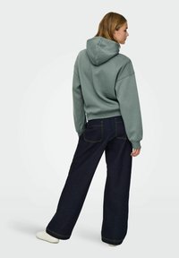 Groene hoodie met een relaxed fit, voorzien van een koord in de capuchon. Gecombineerd met donkerblauwe broek met wijde pijpen, met achterzakken en gele stiksels.