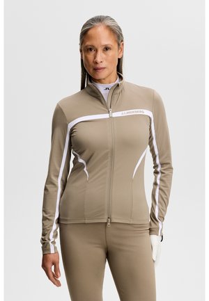 Frau trägt eine enganliegende beige Trainingsjacke mit Reißverschluss, weißen Streifen und passende Hose, steht vor einem schlichten Hintergrund.