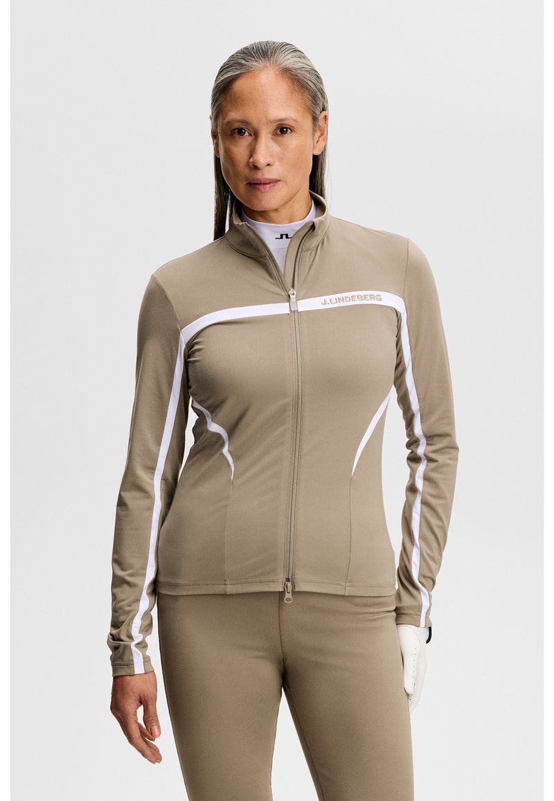 Frau trägt eine enganliegende beige Trainingsjacke mit Reißverschluss, weißen Streifen und passende Hose, steht vor einem schlichten Hintergrund.