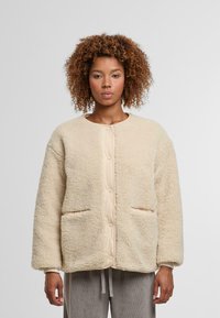 Kobieta z kręconymi włosami w beżowej kurtce szertowej na guziki z przednimi kieszeniami i szarymi prążkowanymi spodniami na jednolitym tle.