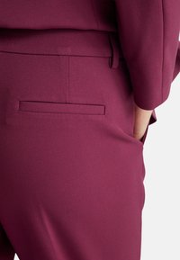 Pantaloni tailored color bordeaux con una superficie liscia, dotati di una tasca posteriore singola senza hardware o decorazioni visibili.