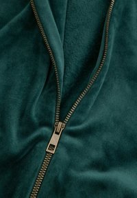 Ciemnozielony materiał o miękkiej, teksturowanej powierzchni z złotą suwakiem częściowo otwartym, odsłaniającym gładki, znajdujący się pod nim materiał.