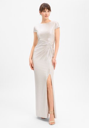 Vrouw draagt een zilverkleurige, korte mouw, vloerlang jurk met zijsamplooien en een hoge split, gecombineerd met sandalen met bandjes, staand tegen een witte achtergrond.