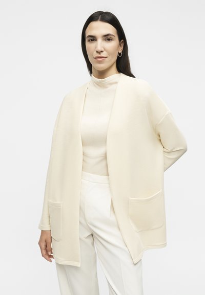 Γυναίκα με σκούρα μαλλιά που φοράει κρεμ ανοιχτή ζακέτα με ρίγες, ανοιχτό ζιβάγκο και λευκό παντελόνι ψηλόμεσο, όρθια με το ένα χέρι πίσω από την πλάτη.