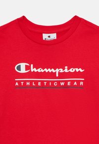 Crvena pamučna majica s okruglim izrezom, sa "Champion" ispisanim bijelim kurzivom i "ATHLETICWEAR" u debelom crnom slovu, smješteno na prednjem dijelu.