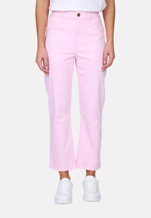 Femme portant un pantalon droit taille haute rose clair, associé à des baskets blanches et un haut blanc rentré, debout devant un fond uni.