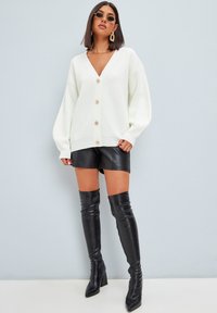 Vit ribbad cardigan med djup V-hals och oversize ärmar, med stora bruna knappar. Kombinerad med svarta lädershorts och knähöga stövlar.