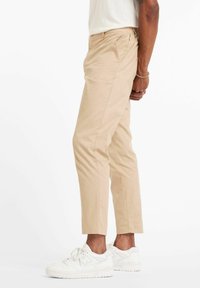 Beige Baumwoll-Chinohosen mit einer schlanken Passform, ausgestattet mit Fronttaschen und einer glatten Textur, kombiniert mit weißen Sneakern.