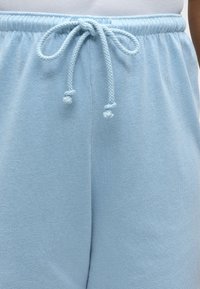 Ανοιχτό μπλε φόρμα με πλεξούδα κορδονιών δεμένη σε φιόγκο στη ελαστική μέση, φορεμένη με λευκή μπλούζα ορατή από πάνω.