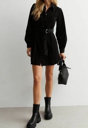 Kobieta ubrana w czarną sukienkę koszulową z paskiem i długimi rękawami, czarne botki za kostkę, trzymająca czarną torebkę na jasnym tle.