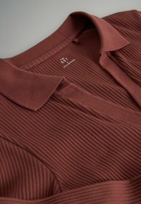 Tricou polo cărnos, bordo, cu guler și cu plastron cu nasturi. Prezintă o textură moale a țesăturii și branding subtil în interiorul gulerului.