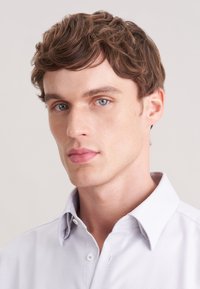 Camicia button-up di un delicato viola chiaro con un motivo testurizzato, dotata di colletto e bottoni bianchi, indossata da una persona con capelli corti e mossi di colore marrone.