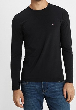 Mężczyzna ubrany w dopasowaną czarną koszulkę z długim rękawem i okrągłym dekoltem z małym logo Tommy Hilfiger na piersi oraz w niebieskie dżinsy.