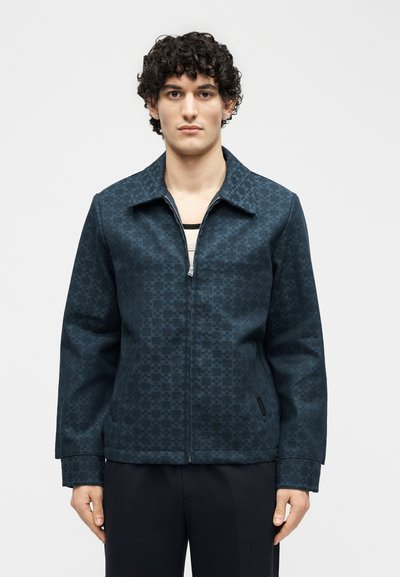 Νεαρός με σγουρά μαλλιά που φοράει σκούρο μπλε μπουφάν με φερμουάρ και μαύρο παντελόνι, στέκεται μπροστά σε απλό φόντο.